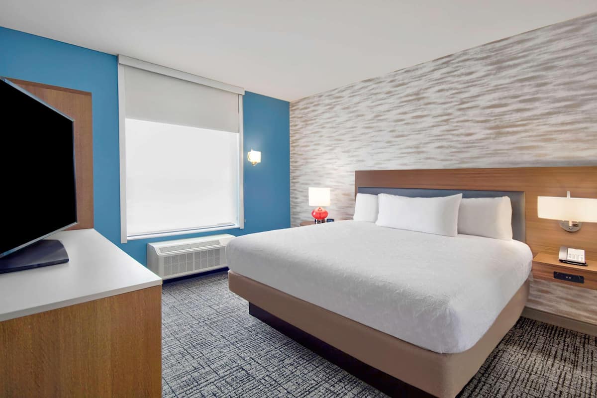 Une chambre d’hôtel dotée d’un grand lit, d’un téléviseur à écran plat, d’une fenêtre avec stores, d’une lampe fixée au mur et d’un mur à texture.