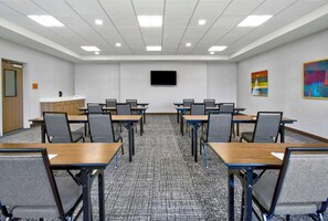 Een vergaderruimte met een wandgemonteerde flatscreen-tv, meerdere tafels en stoelen voor vergaderingen, en een vloerbedekking.