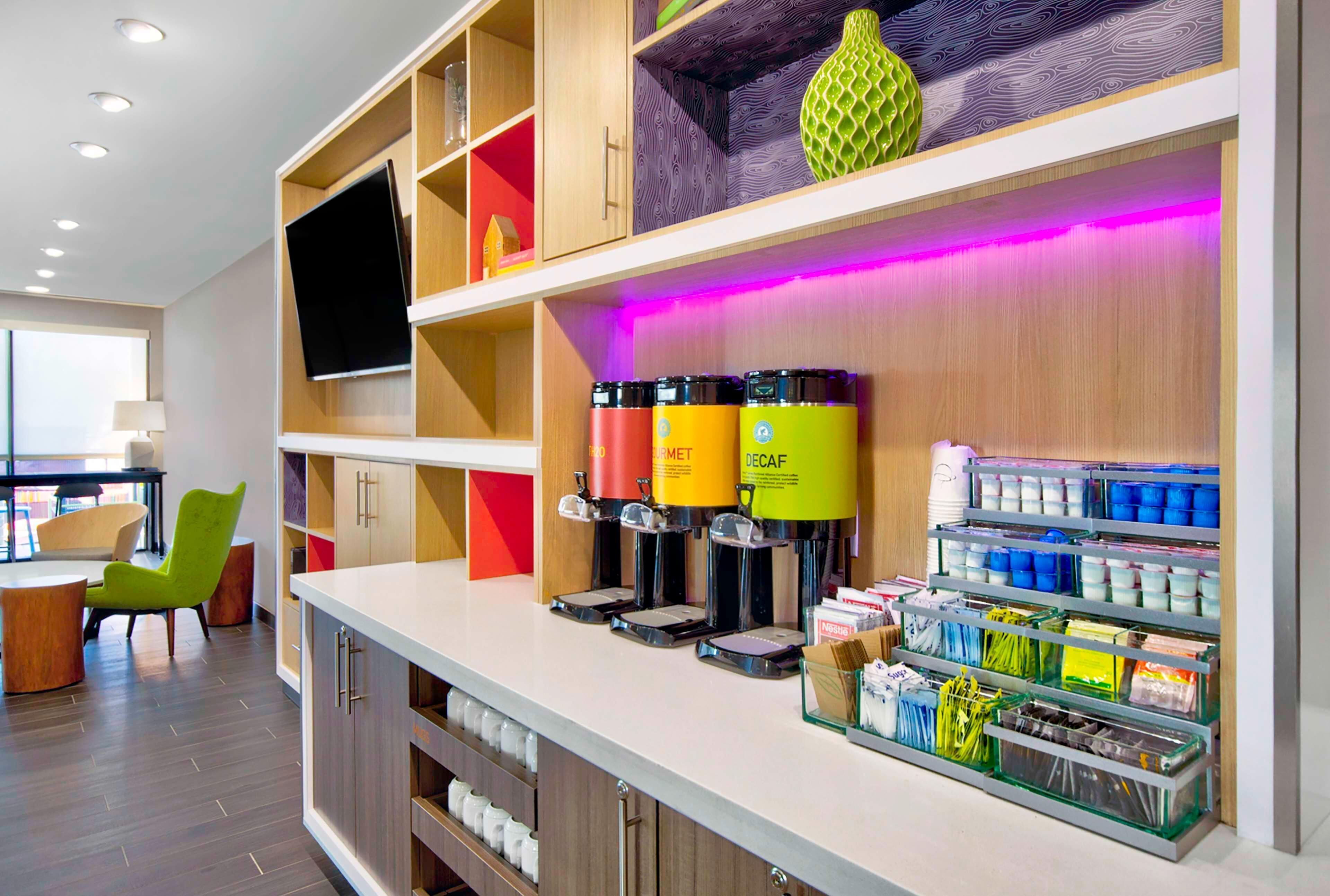 Un moderno vestíbulo de hotel con mostrador de recepción, máquinas de café y estanterías.