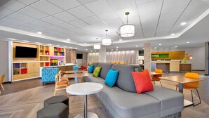Un vestíbul d'hotel modern amb un sofà gris, coixins de colors, una taula rodona blanca i una zona de cuina al fons.