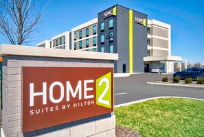 Een hotelschild met 'HOME 2 SUITES BY HILTON' en een gebouw met meerdere ramen en een gele strook.