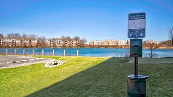 Parque con un cartel que indica limpiar después de los perros, un basurero y una vista de un lago con edificios de apartamentos al fondo.