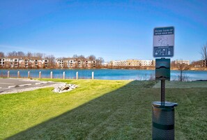 En park med et skilt, der beder om at man rydder op efter hunde, en skraldespand og udsigt til en sø med lejlighedskomplekser i baggrunden.