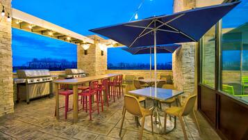 Une terrasse extérieure équipée d’un barbecue, de tables, de chaises et d’un grand parasol.