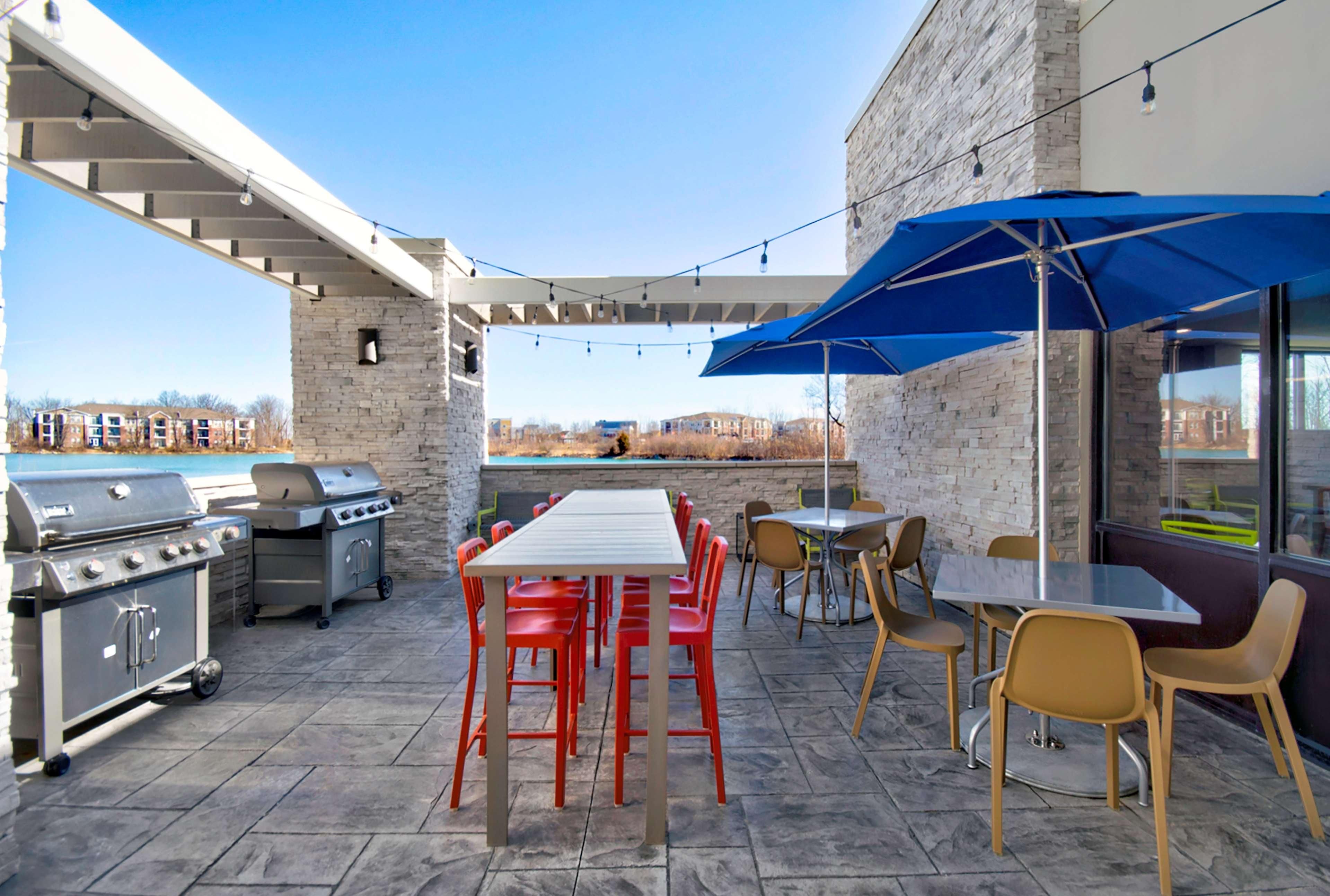 Eine Terrassengruppe im Freien mit Steinboden, Grill, Tisch und Stühlen sowie einem blauen Sonnenschirm.