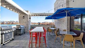 Eine Terrassengruppe im Freien mit Steinboden, Grill, Tisch und Stühlen sowie einem blauen Sonnenschirm.