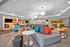 Een moderne hotel lobby met een grijze bank, kleurrijke kussens, een ronde witte tafel en een keuken in de achtergrond.