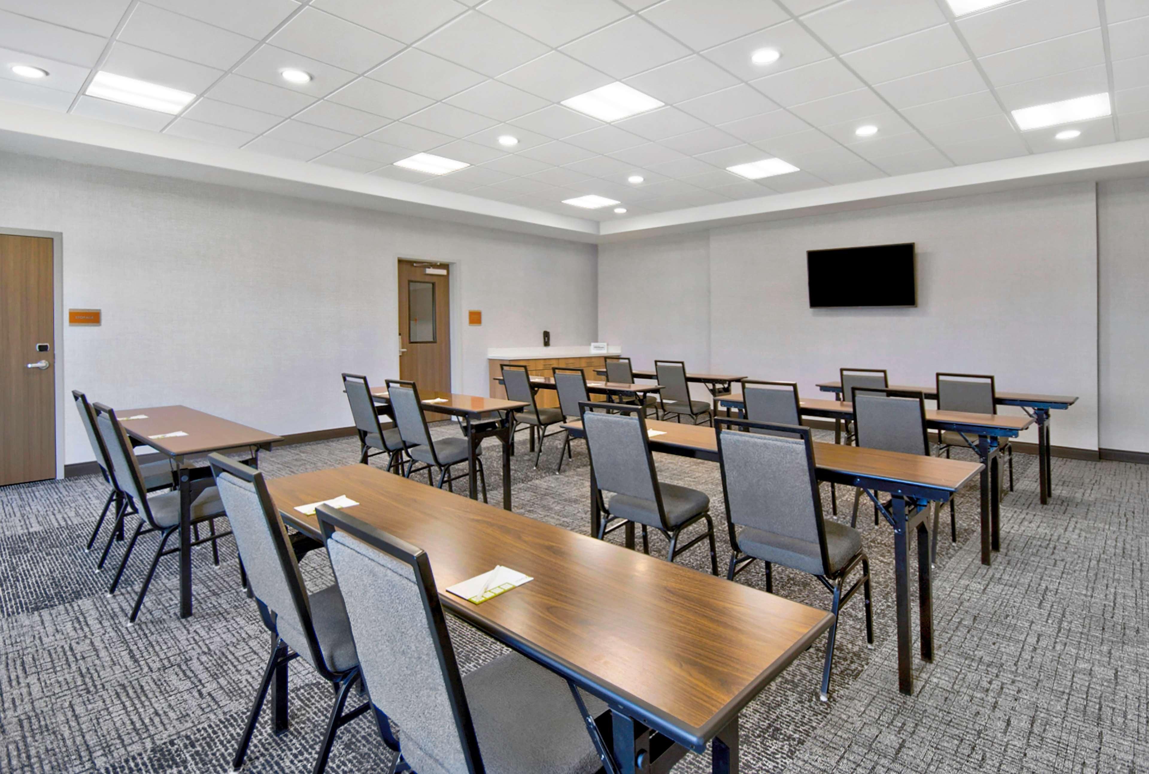 Une salle de conférence dotée de plusieurs tables et chaises, d’un téléviseur à écran plat fixé au mur et d’un revêtement de sol en moquette.