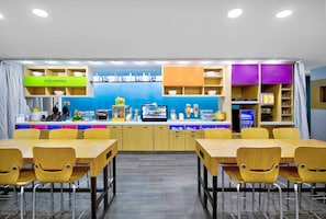 A modern restaurant with a blue wall, wooden tables and chairs, and a counter with various food items. - Home2 Suites by Hilton Whitestown Indianapolis NW (Whitestown)