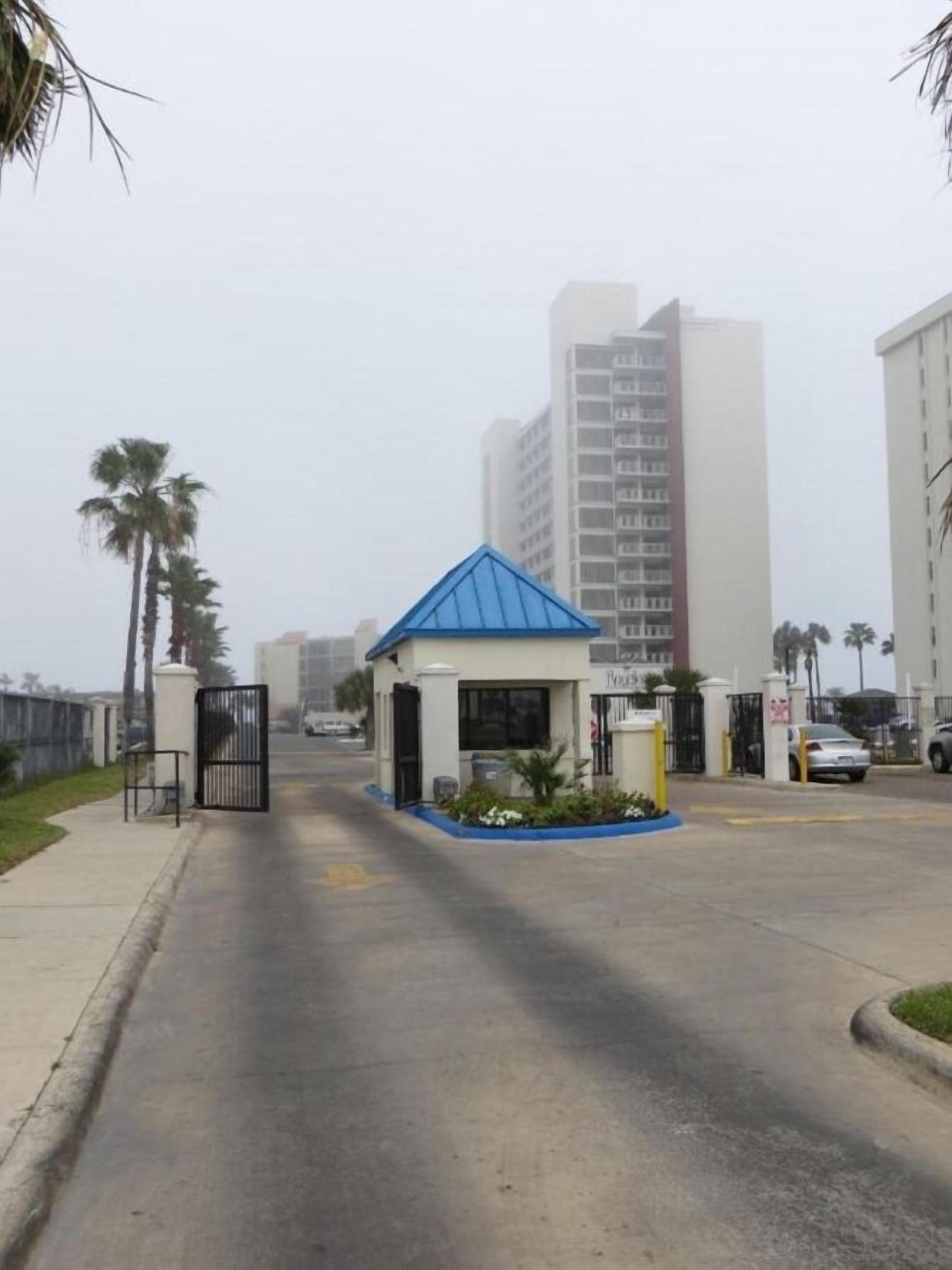 A gated entrance with a small blue-roofed building, palm trees, and tall residential buildings in the background.