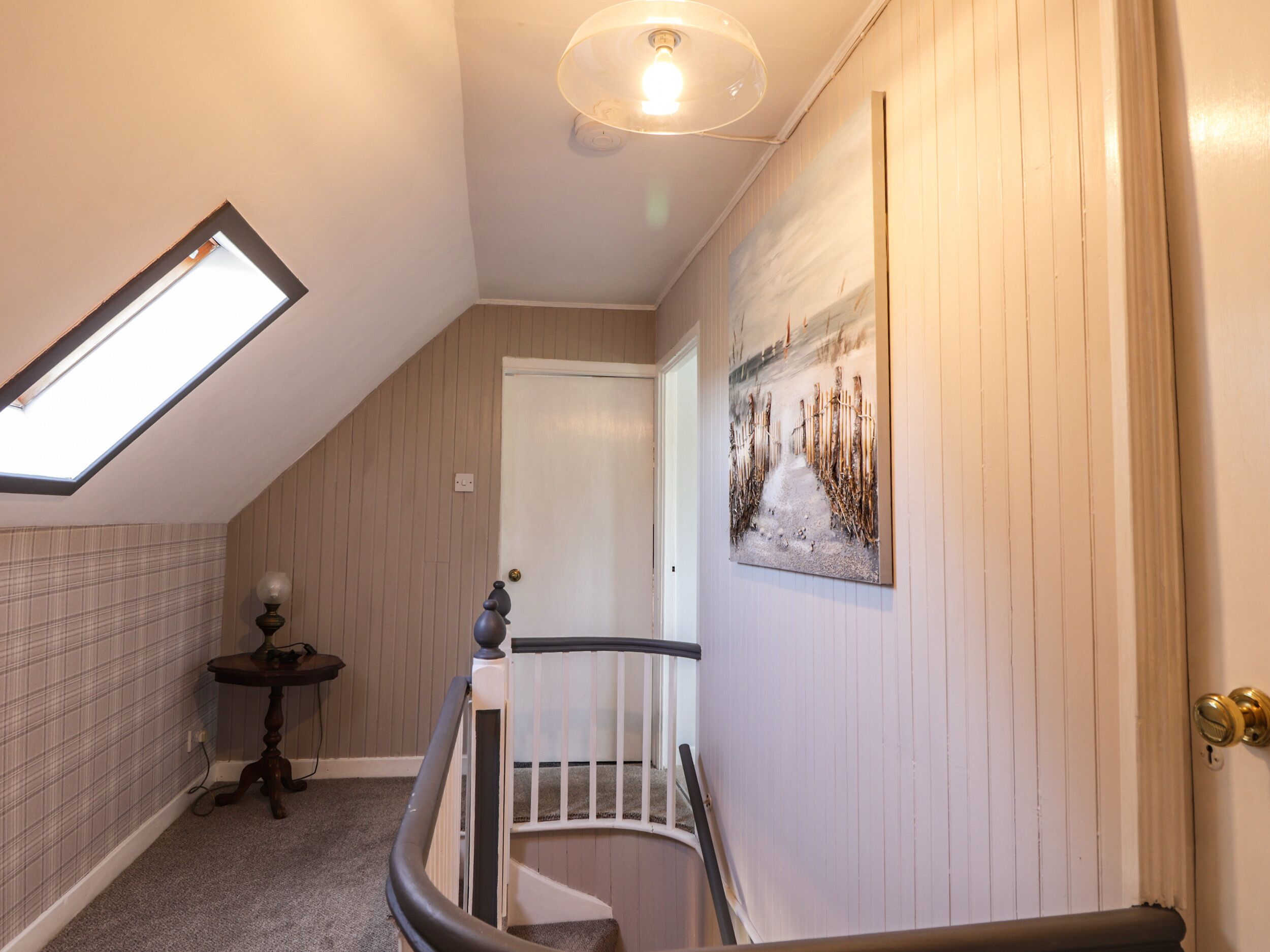 A staircase with a handrail, a small table with a lamp, a painting on the wall, and a skylight.