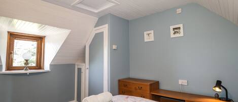 A bedroom with a sloped ceiling, a window with a lamp, a bed with a floral bedspread, a wooden dresser, and a desk with a lamp.