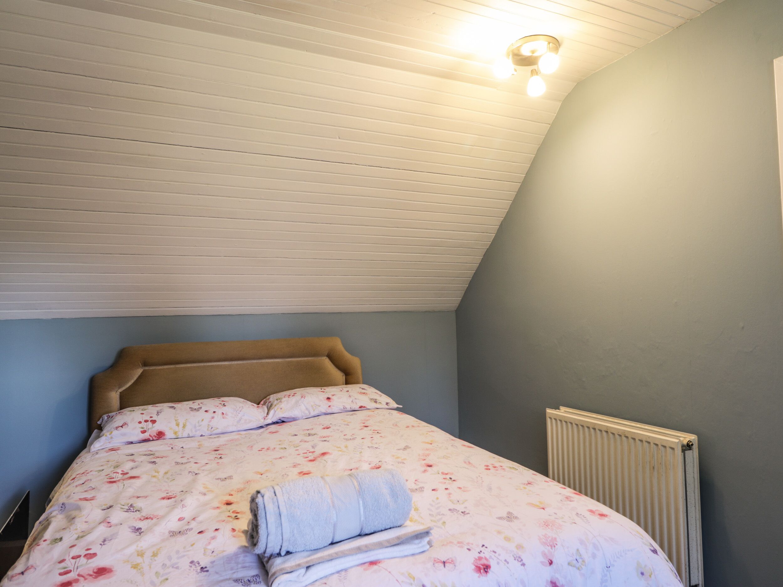 A bedroom with a sloped ceiling, a bed with floral bedding, a rolled towel, and a radiator.