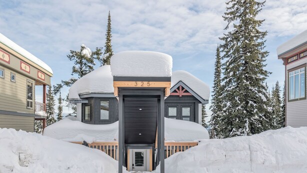 A snow-covered house with a number '325' on the front, surrounded by snowbanks and trees.