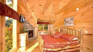 A cozy cabin bedroom with a wooden bed, a fireplace, and a window overlooking a forest. - 3 bed/3 bath luxury cabin with it's own private heated salt water pool! (Sevierville)