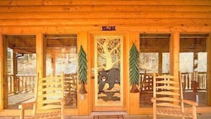 A wooden cabin with a 'Welcome' sign, two wooden chairs, and a door with a bear and tree design. - 3 bed/3 bath luxury cabin with it's own private heated salt water pool! (Sevierville)