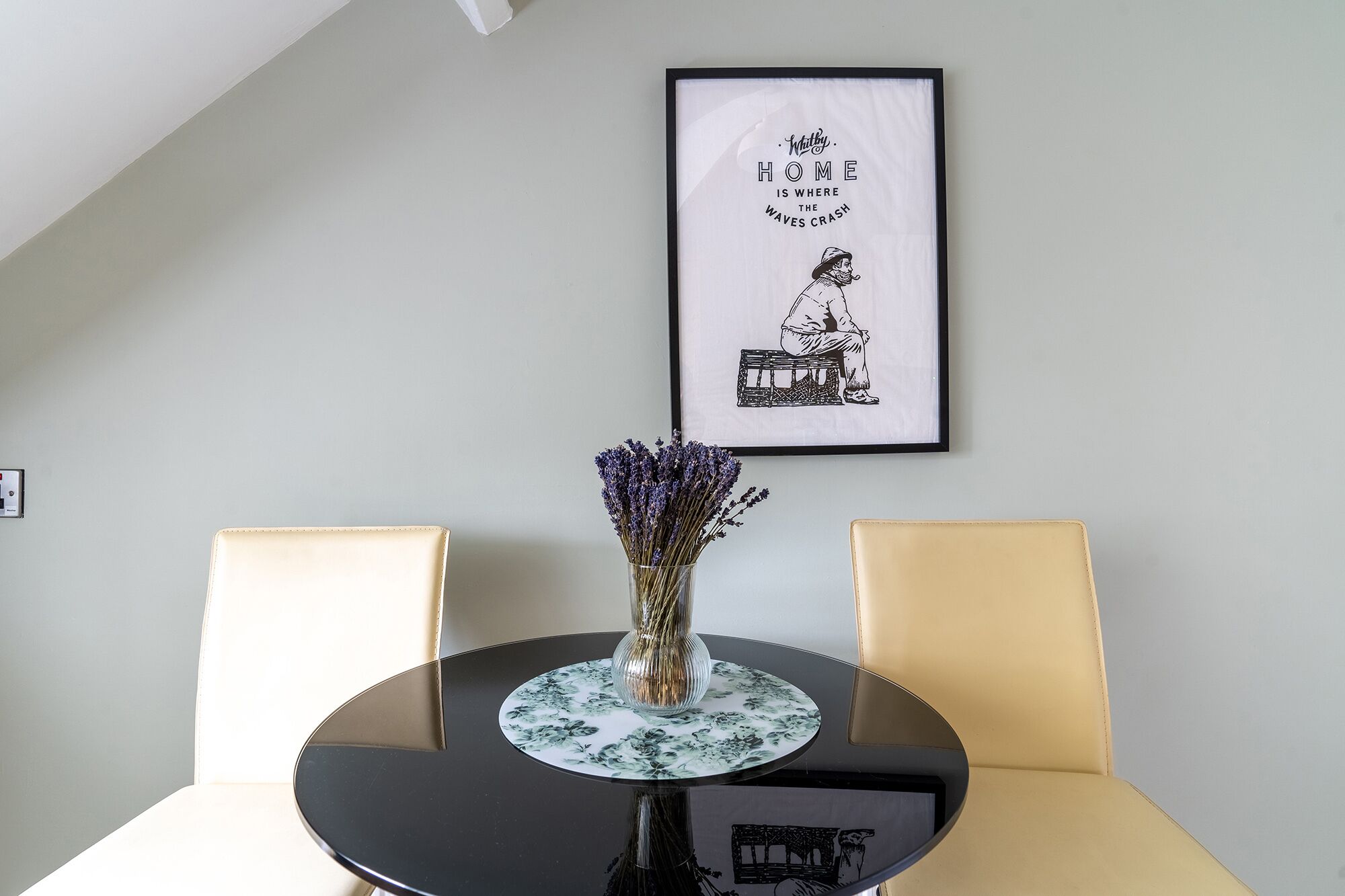 Una mesa de comedor con un jarrón de lavanda, dos sillas y una cita enmarcada en la pared.