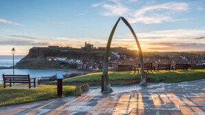 A coastal park with stone benches, a metal arch, and a view of a historic castle on a hill.