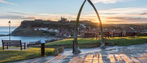 A coastal park with stone benches, a metal arch, and a view of a historic castle on a hill.