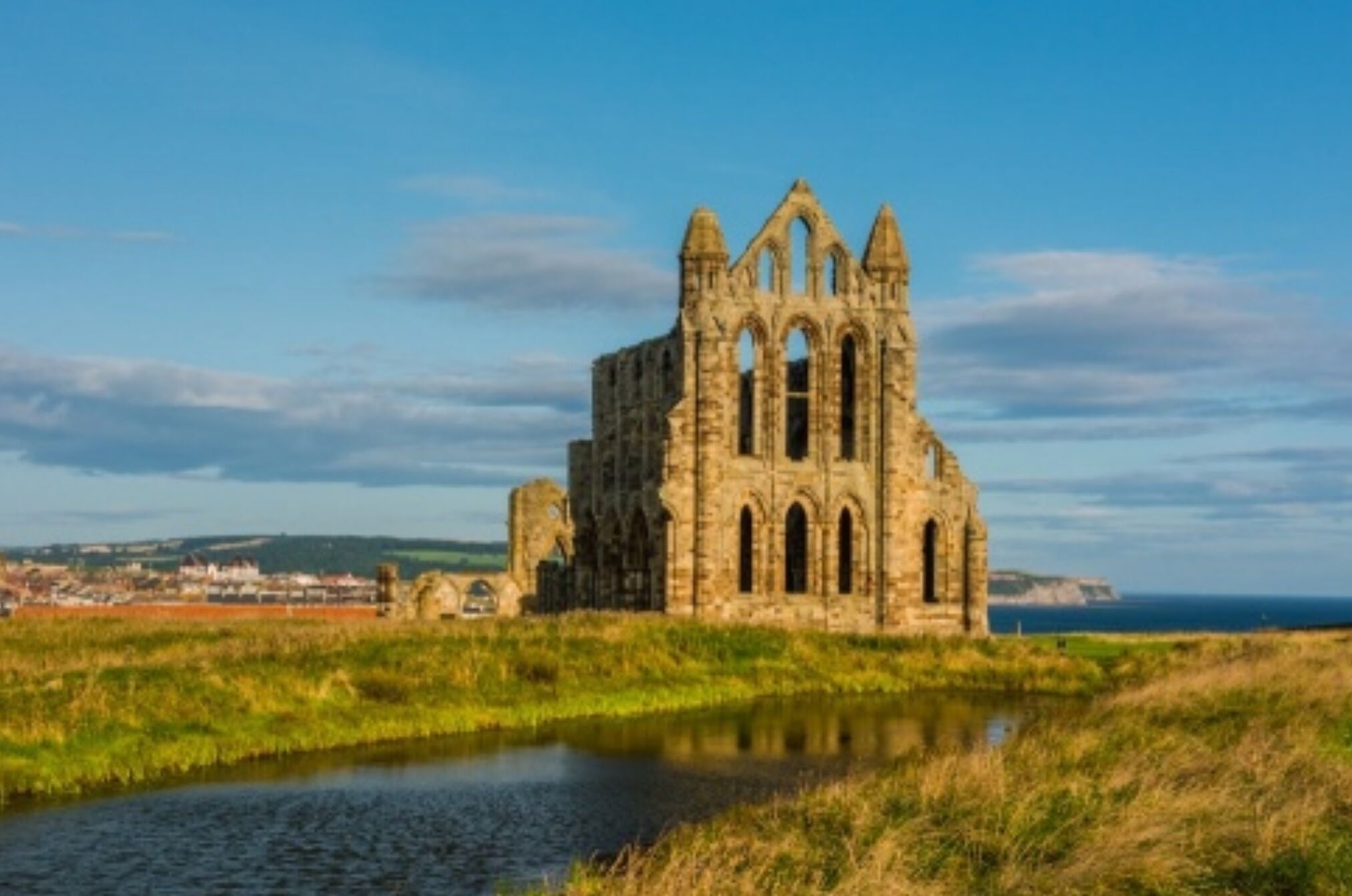 A historic stone building with arched windows and a large central tower, surrounded by a grassy field and a body of water.