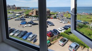 A view from a window overlooking a parking lot with various cars, a building with a flat roof, and a body of water in the distance.