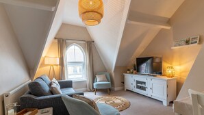 A cozy loft bedroom with a sloped ceiling, a large window with curtains, a TV on a white cabinet, and a comfortable seating area with a sofa and armchairs.