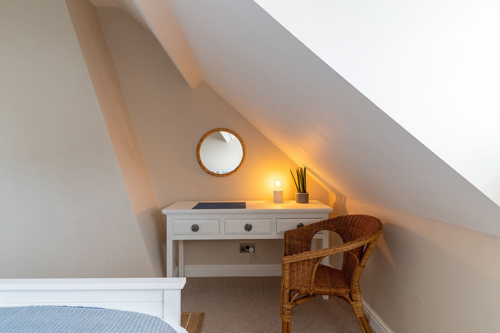 Une chambre avec un plafond en pente, une commode blanche avec des tiroirs, un miroir rond, une petite table avec une lampe et un fauteuil en osier.