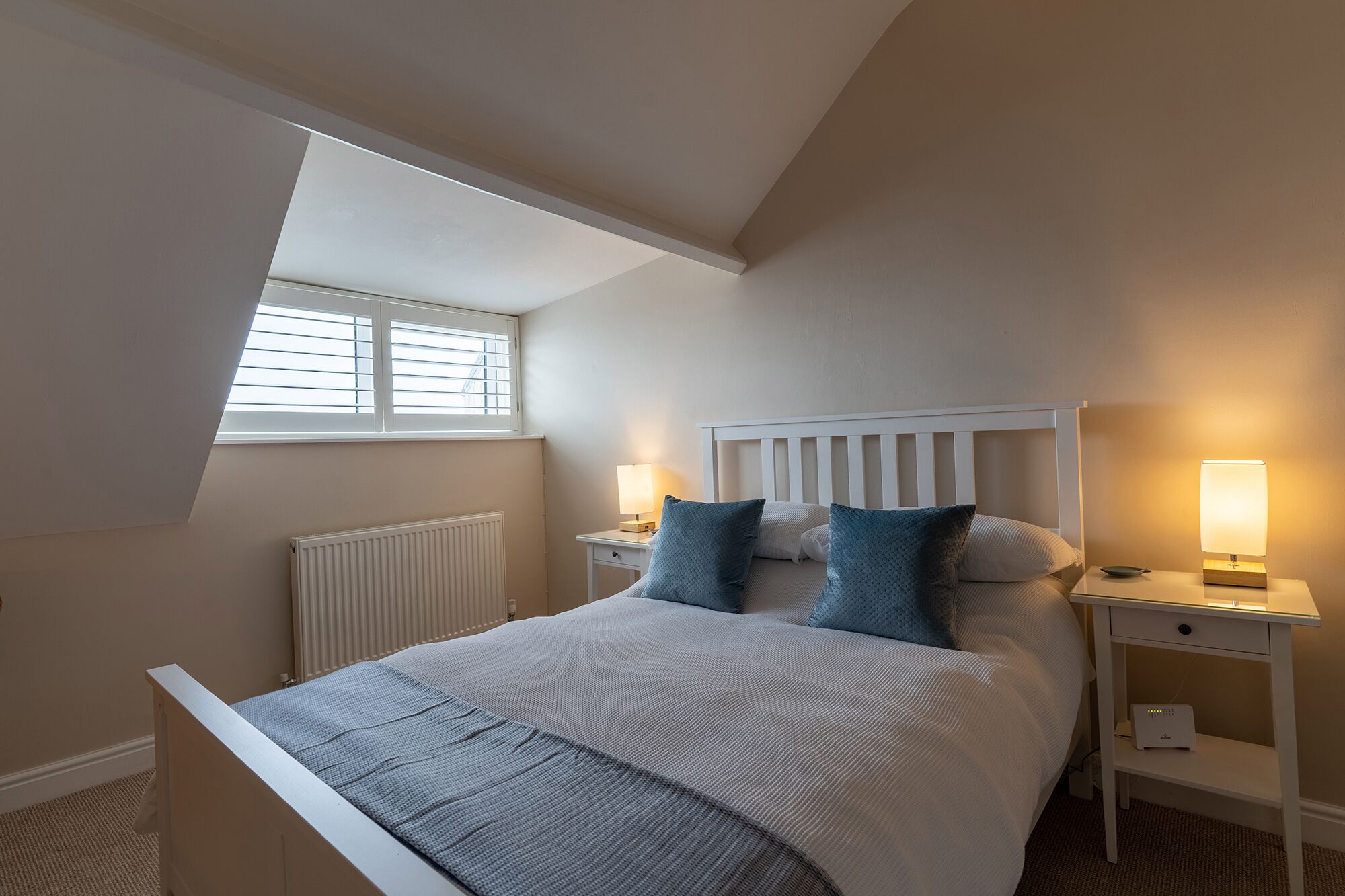 A bedroom with a sloped ceiling, a bed with a white headboard, two bedside tables with lamps, and a window with blinds.