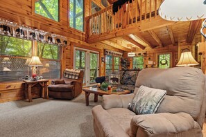Interior acogedor de una cabaña con techo de madera, un ventanal con persianas, un sofá, un sillón y una mesa con lámpara.