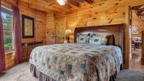 Un dormitorio de cabaña de madera con cama, ventilador de techo, ventana con cortinas y puerta que da a otra habitación.