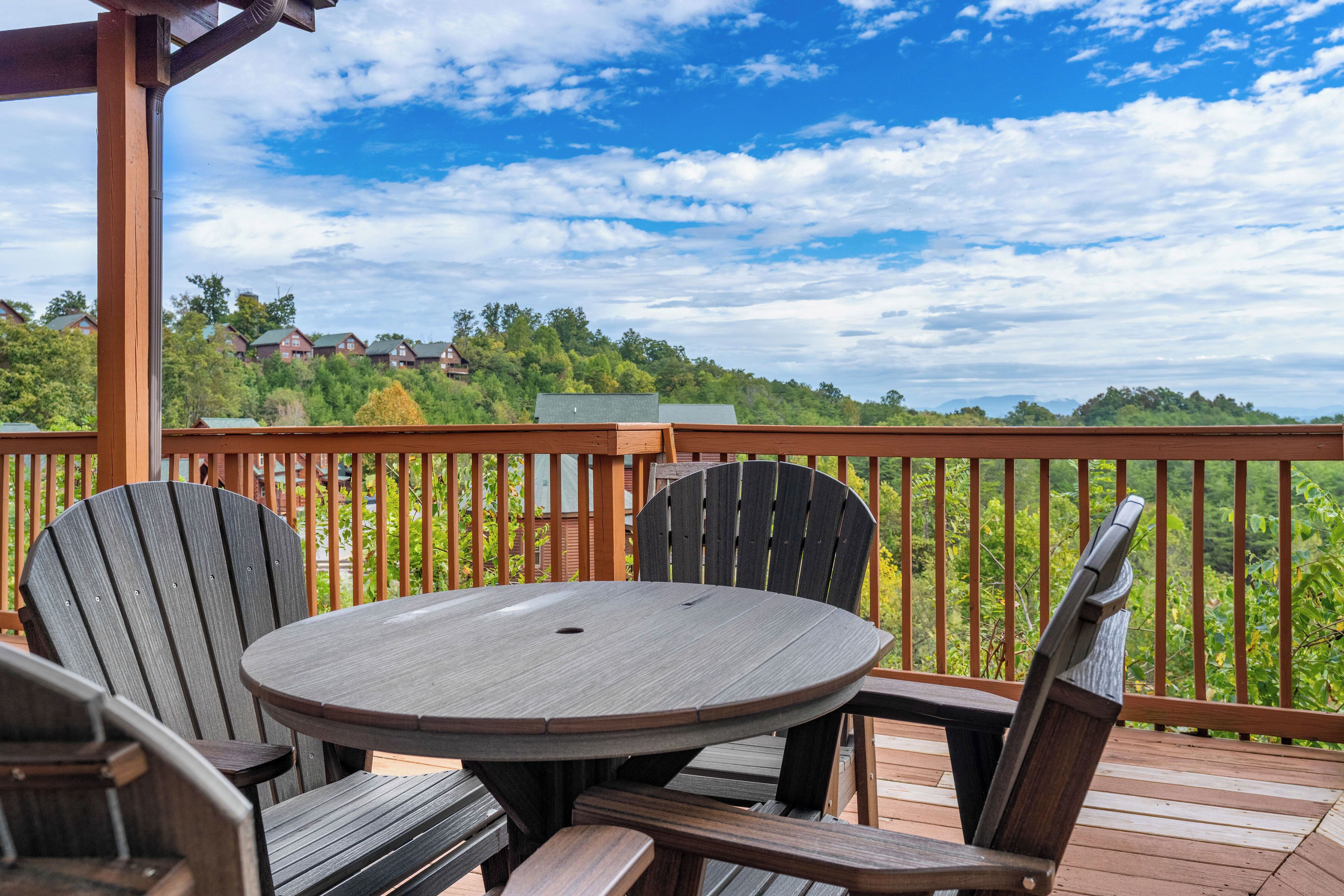 Una terraza de madera con una mesa redonda y sillas Adirondack, con vistas a una zona residencial con casas y árboles.