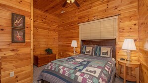 Un dormitorio de cabaña de madera con cama, dos mesitas de noche, ventilador de techo y colcha.