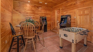 Una habitación con piso de madera, una mesa de fútbol de mesa, dos juegos de arcade y un comedor con sillas y una mesa.