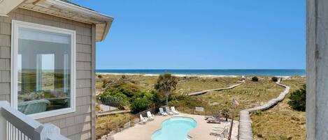 A pool with lounge chairs, a wooden deck, and a view of the beach and ocean.