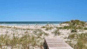 Ein Holzsteg führt zu einem Strand mit Dünen und dem Meer im Hintergrund.
