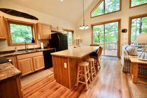 A spacious kitchen with wooden cabinets, a large island with stools, and a view of the outdoors through large windows. - South Turtle Lake! Private Dock! Pet-friendly, Canoe Included!August dates open! (Winchester)