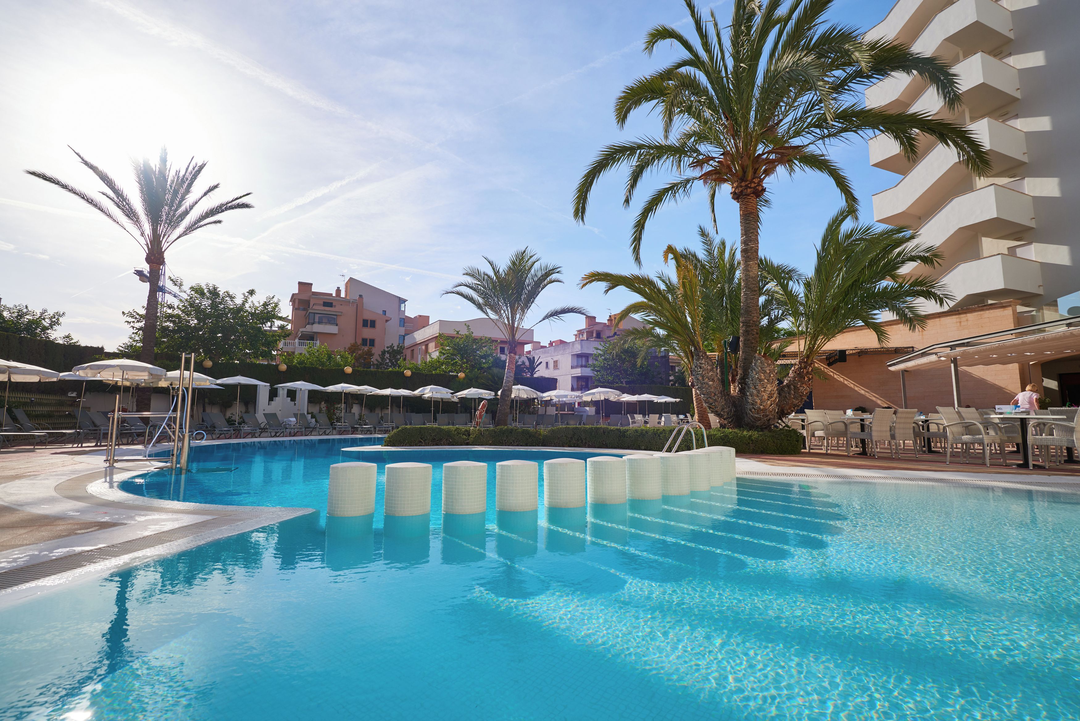 Une piscine entourée de palmiers et d’immeubles, avec des chaises longues et des parasols.