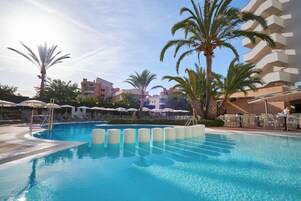 Piscina con sillas de descanso y sombrillas, rodeada de palmeras y edificios.