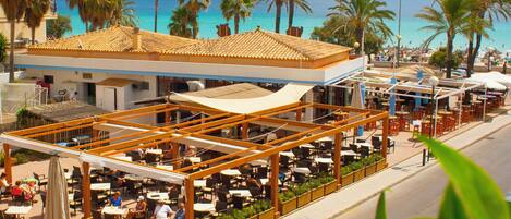 An outdoor café with tables and chairs, overlooking a beach with palm trees and the sea.