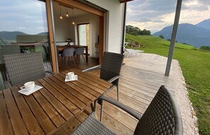 Une terrasse en bois, agrémentée de chaises et d’une table en osier, offre une vue imprenable sur un paysage verdoyant, avec des montagnes en toile de fond.