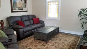 A living room with a dark leather sofa, a wooden coffee table, a patterned rug, a large window with blinds, and a potted plant. - Hatch Life in Steinhatchee Landing (Steinhatchee)