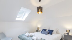 Un dormitorio moderno con tragaluz, cama con ropa de cama blanca y azul, dos mesitas de noche con lámparas y una silla gris.