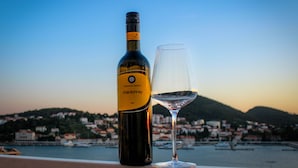 Een fles chardonnay en een wijnglas op een balkon met uitzicht op een kustplaatsje en heuvels.