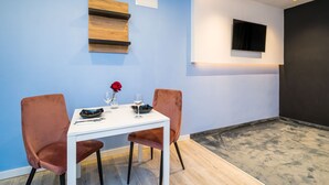 Een eethoek met een witte tafel, twee fluweelzachte stoelen, een rozenvaas en een flatscreen televisie aan de muur.