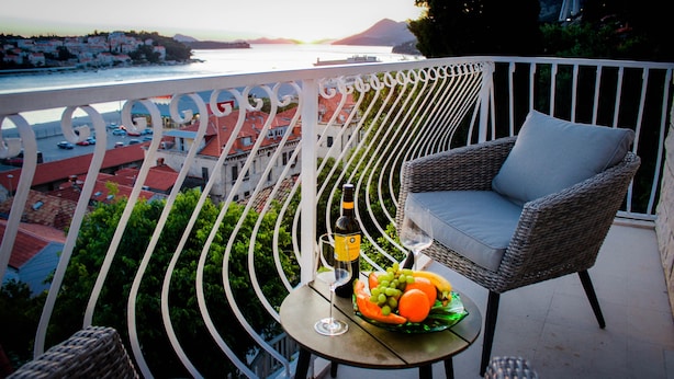 Een balkon met een rieten stoel, een tafel met een fles en fruit, en uitzicht op een kustplaats.