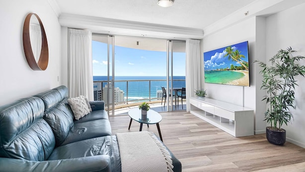 A modern living room with a leather sofa, a small coffee table, a flat-screen TV, and a large window with a view of the ocean. - Chevron Renaissance Ocean View (Surfers Paradise)