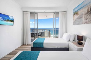 A hotel room with two beds, a balcony with a view of the ocean, a painting of a beach scene, and a television on the wall.