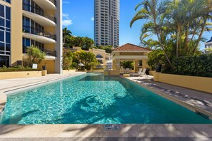 A swimming pool with clear water, surrounded by a building with balconies and a tall skyscraper in the background.