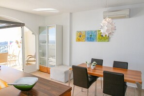 Un comedor moderno con una mesa de madera, sillas negras y un panel de vidrio. Se observan obras de arte en la pared y una unidad de aire acondicionado.
