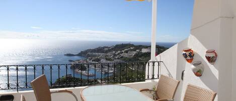 Ein Balkon mit Korbstühlen und einem Tisch, der Blick auf die Küste mit Gebäuden und dem Meer freigibt.
